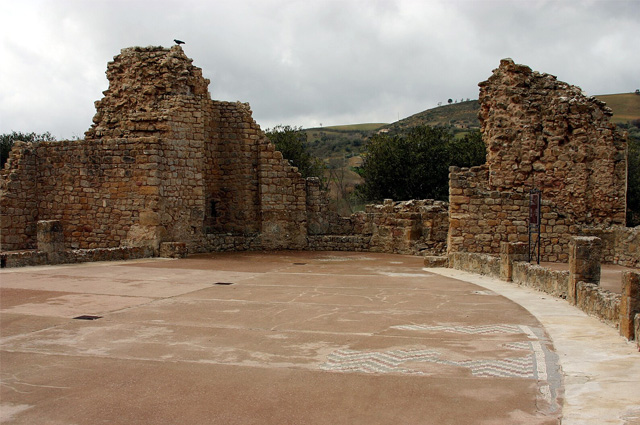 The remains of two brick walls at the back of a smooth floor covered in large slabs of stone with hills in the background.