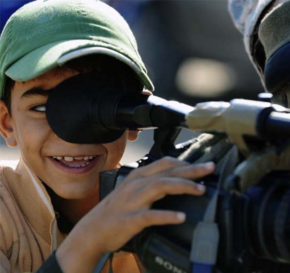 Young boy in a green baseball cap peers through a video camera viewfinder.
