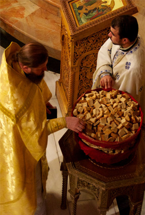 A priest in a yellow robe chooses a triangle of bread from a basket while another priest in a white top with blue embroidery holds the red basket.